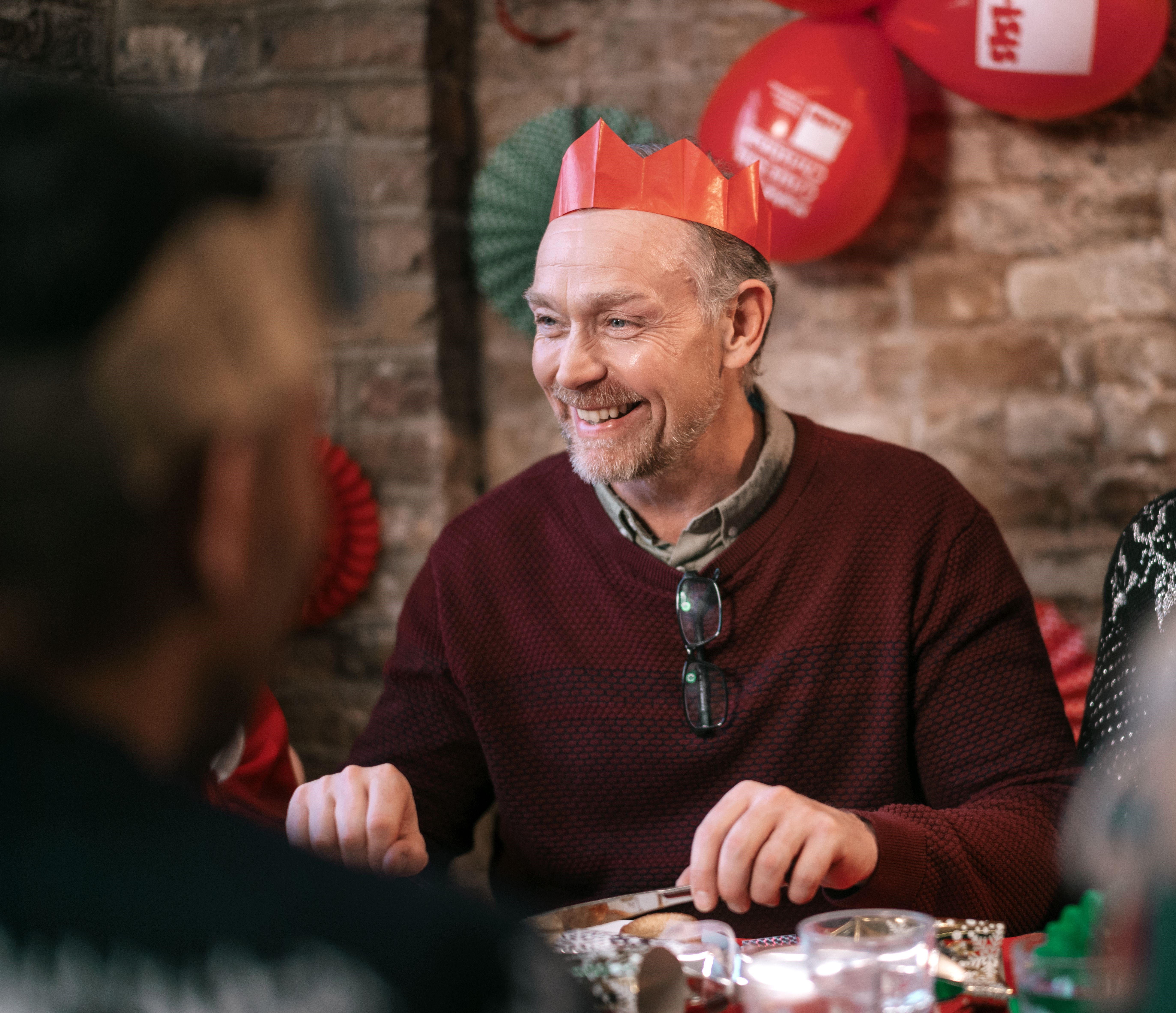 Simon is smiling broadly while wearing a paper crown from a Christmas cracker. Behind him there are Crisis branded balloons and Christmas decorations.