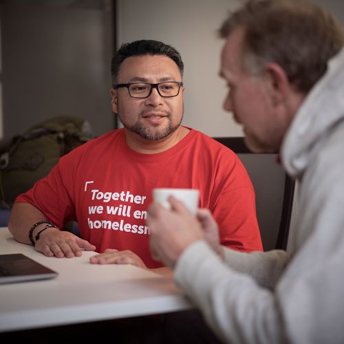 Simon is sat with a Crisis staff member. Simon is drinking a cup of tea and the staff member is listening to him.